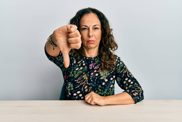 Beautiful middle age woman wearing casual clothes sitting on the table looking unhappy and angry showing rejection and negative with thumbs down gesture. bad expression.  - Photo, Image