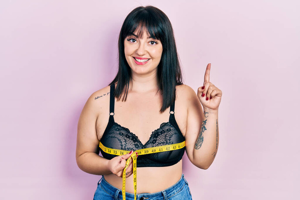 Young hispanic woman using tape measure measuring breast surprised with an idea or question pointing finger with happy face, number one  - Photo, Image