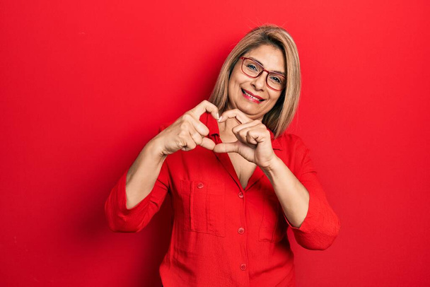 Middelbare leeftijd Spaanse vrouw dragen casual kleding en glazen glimlachen in liefde tonen hart symbool en vorm met de handen. romantisch concept.  - Foto, afbeelding
