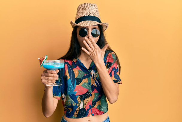 Junge hispanische Frau im Sommer-Stil trinkt tropischen Cocktail mit Alkohol bedeckt Mund mit der Hand, schockiert und Angst vor Fehler. Überraschte Miene  - Foto, Bild