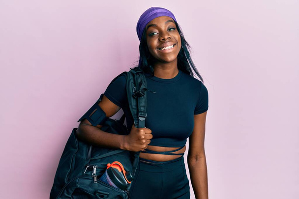 Молодая африканская американка с спортивной сумкой выглядит позитивно и счастливо стоя и улыбаясь с уверенной улыбкой, показывая зубы  - Фото, изображение