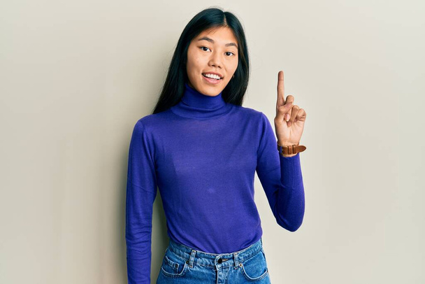 Jeune femme chinoise portant des vêtements décontractés souriant avec une idée ou une question pointant du doigt avec un visage heureux, numéro un  - Photo, image