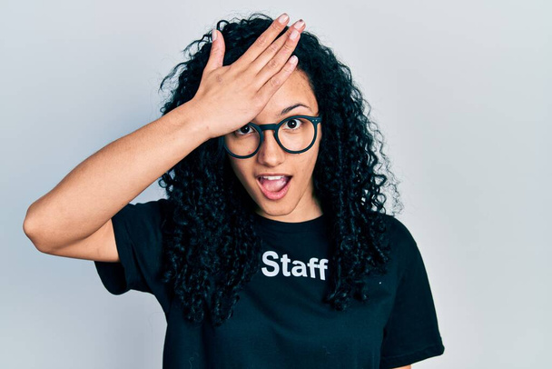 Young hispanic woman with curly hair wearing staff t shirt surprised with hand on head for mistake, remember error. forgot, bad memory concept.  - Photo, Image