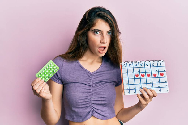 Junge kaukasische Frau mit Antibabypille und Menstruationskalender im Schockgesicht, sieht skeptisch und sarkastisch aus, überrascht mit offenem Mund  - Foto, Bild