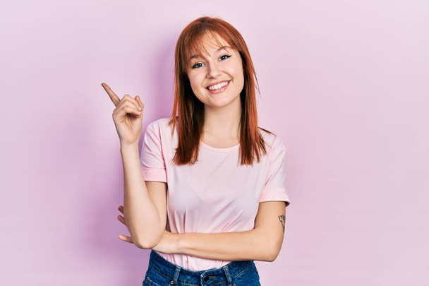 Rossa giovane donna indossa casual t shirt rosa con un grande sorriso sul viso, puntando con mano e dito verso il lato guardando la fotocamera.  - Foto, immagini