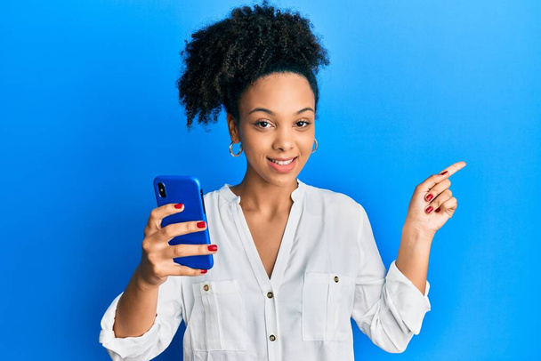 Młoda afrykańska dziewczyna za pomocą smartfona uśmiechnięty szczęśliwy wskazując z ręki i palca na bok  - Zdjęcie, obraz