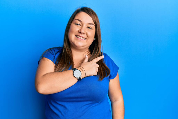 Красивая брюнетка плюс размер женщина в повседневной синей футболке весело с улыбкой на лице указывая рукой и пальцем в сторону со счастливым и естественным выражением лица  - Фото, изображение