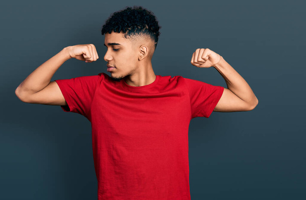 Jovem afro-americano vestindo camiseta vermelha casual mostrando os músculos dos braços sorrindo orgulhoso. conceito de aptidão.  - Foto, Imagem
