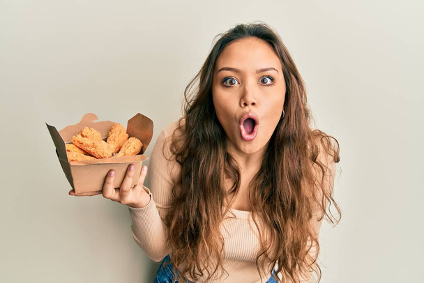 Mladý hispánec dívka jíst kuřecí křidélka strach a ohromen s otevřenými ústy pro překvapení, nevěřící tvář  - Fotografie, Obrázek