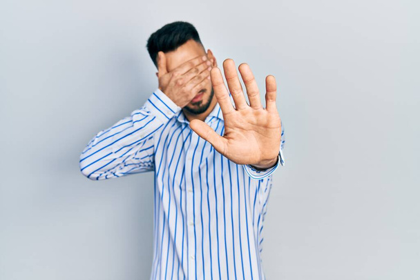 Jonge Latijns-Amerikaanse man met baard draagt casual gestreept shirt dat ogen bedekt met handen en doet stop gebaar met droevige en angstexpressie. beschaamd en negatief concept.  - Foto, afbeelding