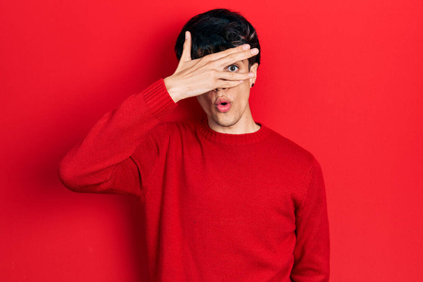 Jóképű, hipszter fiatalember piros téli pulóvert visel, sokk borítja az arcát és a szemét, kezével néz keresztül az ujjain, fél  - Fotó, kép