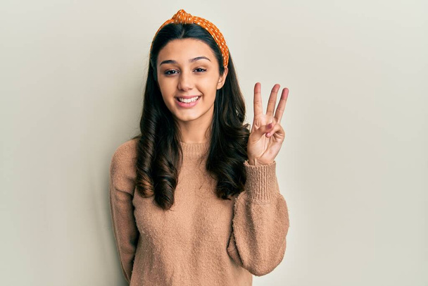 Молодая латиноамериканка, одетая в повседневную одежду и показывающая пальцем номер три, улыбаясь уверенно и счастливо.  - Фото, изображение