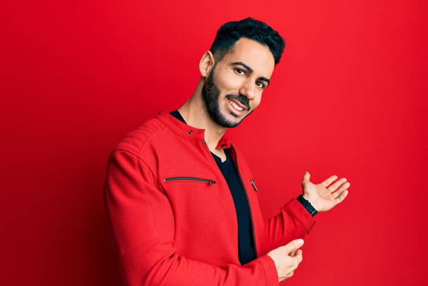 Jonge Spaanse man draagt rode leren jas en nodigt uit om binnen te komen lachend natuurlijk met open hand  - Foto, afbeelding