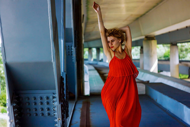 Όμορφη Λάμψη Ξανθιά γυναίκα με σγουρά μαλλιά σε κόκκινο φόρεμα που χορεύουν κάτω από τη γέφυρα. - Φωτογραφία, εικόνα