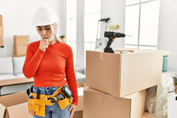 Middle age grey-haired woman wearing hardhat standing at new home feeling unwell and coughing as symptom for cold or bronchitis. health care concept.  - Photo, Image