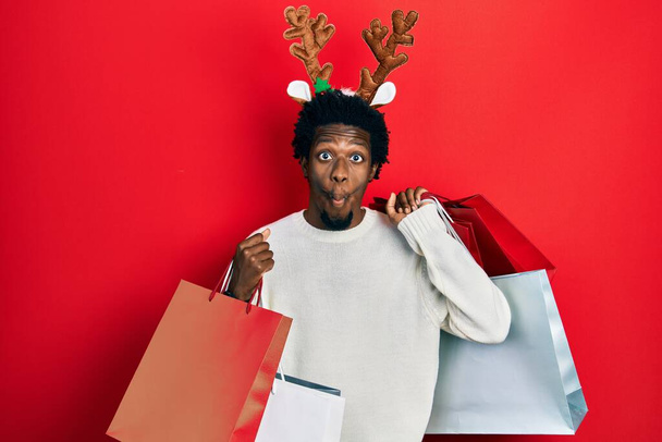 Jonge Afro-Amerikaanse man draagt herten kerst hoed met boodschappentassen maken vis gezicht met mond en squinting ogen, gek en komisch.  - Foto, afbeelding