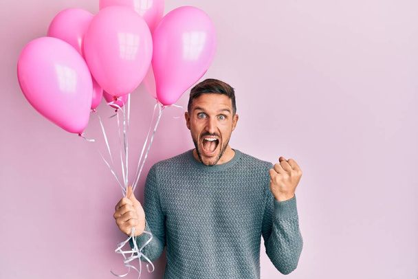 Homem bonito com barba segurando balões cor-de-rosa gritando orgulhoso, celebrando vitória e sucesso muito animado com braços levantados  - Foto, Imagem