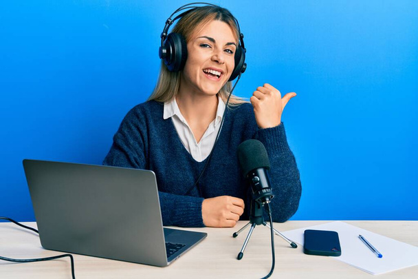 jonge Kaukasische vrouw werken op radio studio duim wijzen naar de zijkant glimlachen blij met open mond  - Foto, afbeelding