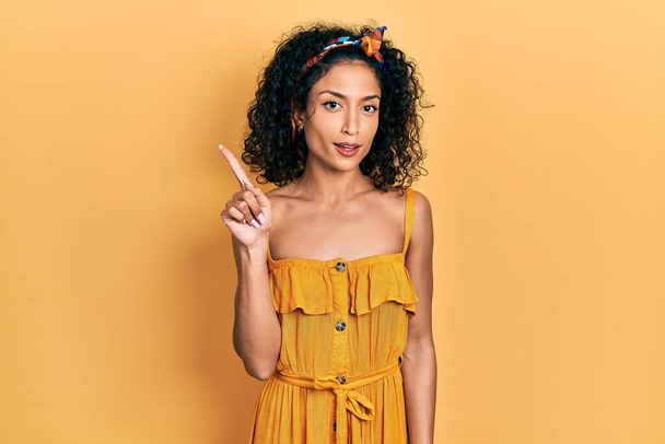 Joven chica latina con vestido de verano mostrando y señalando hacia arriba con el dedo número uno mientras sonríe confiado y feliz.  - Foto, Imagen