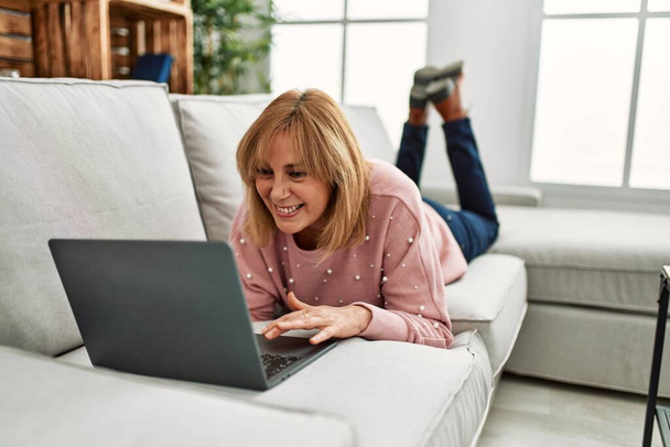 Blondynka w średnim wieku korzystająca z laptopa leżącego na kanapie w domu. - Zdjęcie, obraz