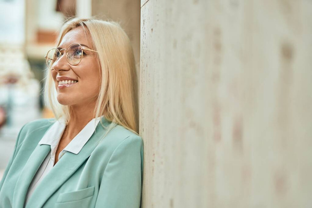 Mladý blondýnka businesswoman usměvavý šťastný stojící ve městě - Fotografie, Obrázek