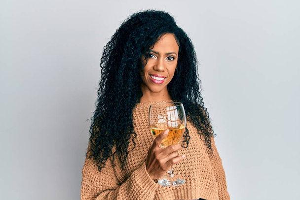 Orta yaşlı Afro-Amerikalı bir kadın bir bardak beyaz şarap içiyor. Pozitif görünüyor ve gülümsüyor. Kendine güvenen bir gülümsemeyle dişlerini gösteriyor.  - Fotoğraf, Görsel