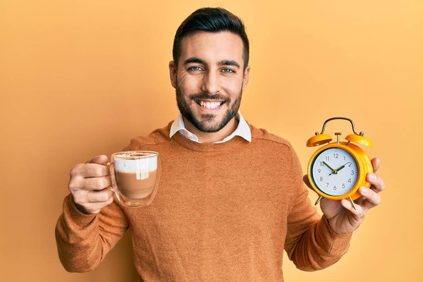Fiatal spanyol férfi kezében kávé és ébresztőóra mosolyog egy boldog és hűvös mosollyal az arcán. fogak megjelenítése.  - Fotó, kép