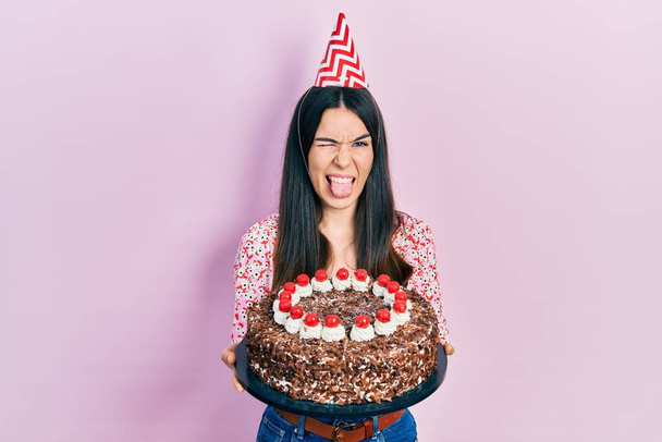 Junge brünette Frau feiert Geburtstag mit großer Schokoladenkuchen, die Zunge herausstreckt, glücklich mit lustigem Ausdruck.  - Foto, Bild