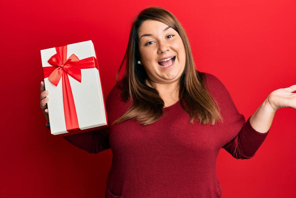 Beautiful brunette plus size woman holding gift celebrating achievement with happy smile and winner expression with raised hand  - Photo, Image