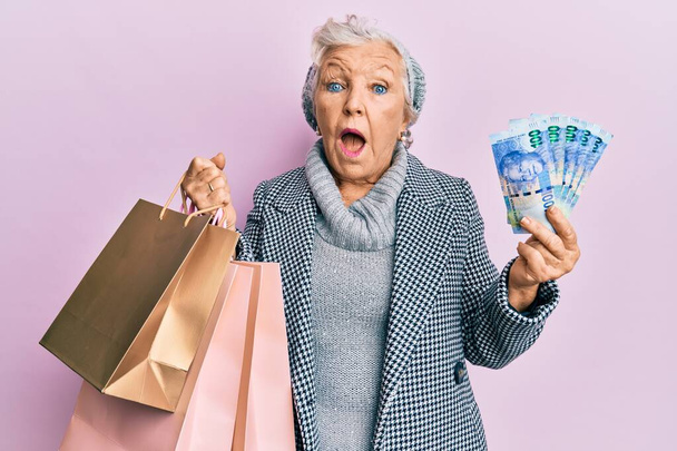 Femme âgée aux cheveux gris tenant des sacs à provisions et des billets de banque en rands d'Afrique du Sud effrayée et choquée par la surprise et l'expression étonnante, la peur et le visage excité.  - Photo, image