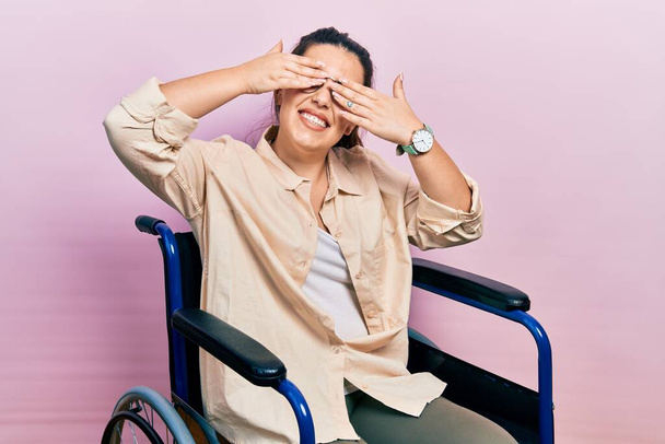 Young hispanic woman sitting on wheelchair covering eyes with hands smiling cheerful and funny. blind concept.  - Fotó, kép