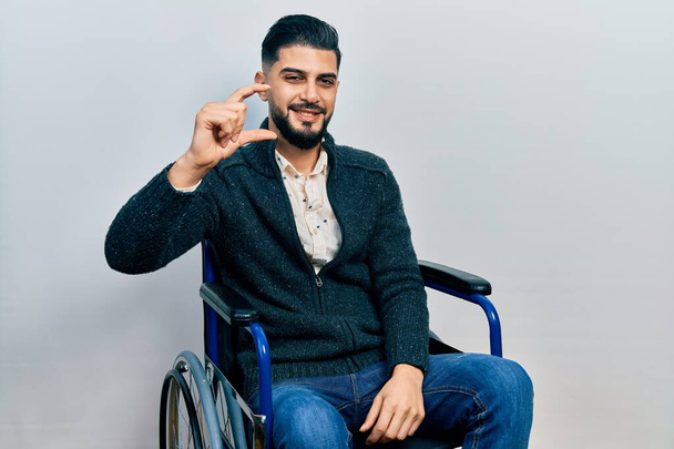 Pohledný muž s vousy sedící na invalidním vozíku, usmívající se a sebevědomý, gestikuluje rukou a malými prsty se dívá do kamery. koncepce ukazatele.  - Fotografie, Obrázek