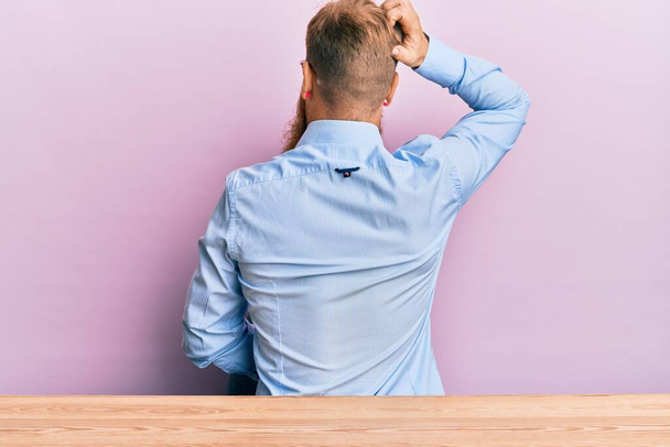 Mladý irský zrzavý muž v obchodní košili a kravatě sedí na stole pozpátku a přemýšlí o pochybách s rukou na hlavě  - Fotografie, Obrázek
