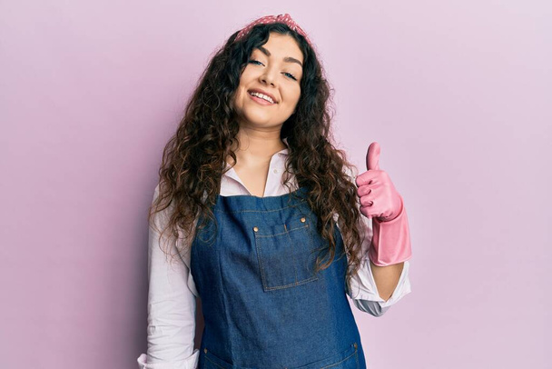 Young brunette woman with curly hair wearing cleaner apron and gloves doing happy thumbs up gesture with hand. approving expression looking at the camera showing success.  - Foto, Imagem