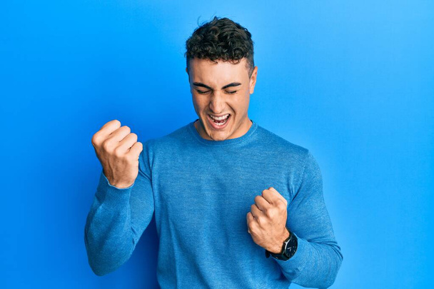 Hispanic young man wearing casual winter sweater celebrating surprised and amazed for success with arms raised and eyes closed. winner concept.  - Fotografie, Obrázek