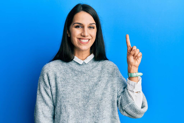 Genç İspanyol kadın gündelik kıyafetler giyip bir numarayı gösterip işaret ederken kendinden emin ve mutlu bir şekilde gülümsüyor..  - Fotoğraf, Görsel