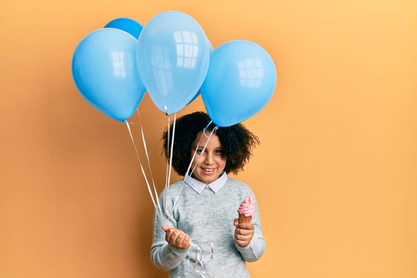 Νεαρό κοριτσάκι με αφρο μαλλιά κρατώντας παγωτό και μπλε μπαλόνια χαμογελώντας και γελώντας δυνατά γιατί αστείο τρελό αστείο.  - Φωτογραφία, εικόνα