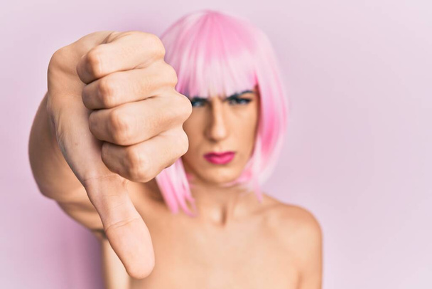 Młody mężczyzna ubrany w kobietę tworzą sobie różową perukę wygląda nieszczęśliwy i zły pokazując odrzucenie i negatywne z kciukami w dół gest. złe wyrażenie.  - Zdjęcie, obraz