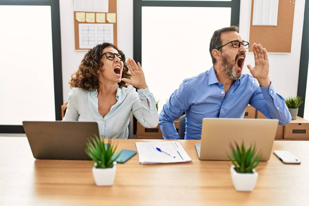 Middle age hispanic woman and man sitting with laptop at the office shouting and screaming loud to side with hand on mouth. communication concept.  - Photo, Image