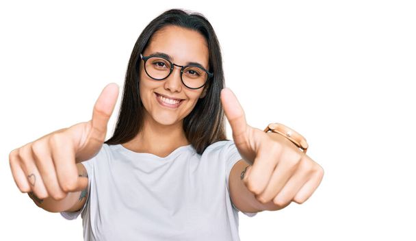 若いヒスパニック系の女性は、手で肯定的なジェスチャーを行うことを承認カジュアルな白いTシャツを着て,親指アップ笑顔と成功のために幸せ.勝者のジェスチャー.  - 写真・画像
