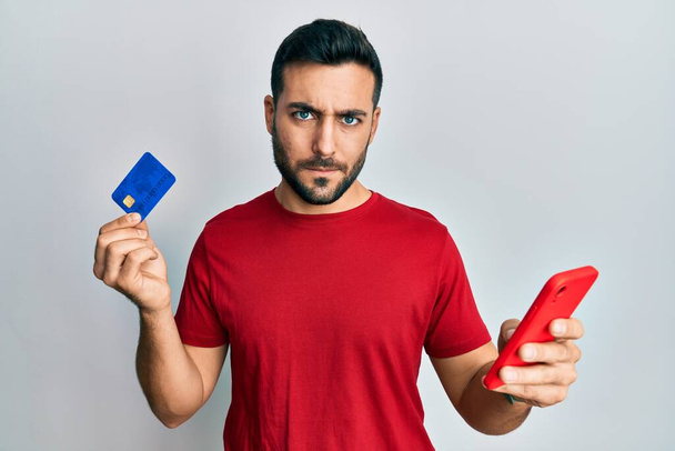 Joven hombre hispano sosteniendo teléfono inteligente y tarjeta de crédito escéptico y nervioso, frunciendo el ceño molesto debido a un problema. persona negativa.  - Foto, Imagen