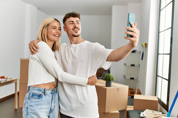 Νεαρό καυκάσιο ζευγάρι χαμογελά χαρούμενος κάνοντας selfie από το smartphone στο νέο σπίτι. - Φωτογραφία, εικόνα