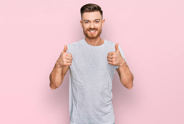 Mladý rusovlasý muž v neformální šedé tričko s nápisem úspěch dělá pozitivní gesto rukou, palce nahoru s úsměvem a šťastný. veselý výraz a gesto vítěze.  - Fotografie, Obrázek