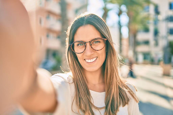 Молода біла жінка, посміхаючись щаслива, робить селфі біля камери в місті
. - Фото, зображення