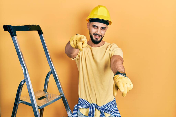 Красивый мужчина с бородой по строительной лестнице в каске указывает на тебя и камеру пальцами, улыбаясь позитивно и весело  - Фото, изображение