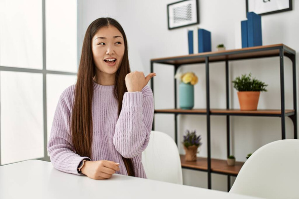 Young chinese girl wearing casual clothes sitting on the table at home smiling with happy face looking and pointing to the side with thumb up.  - Valokuva, kuva