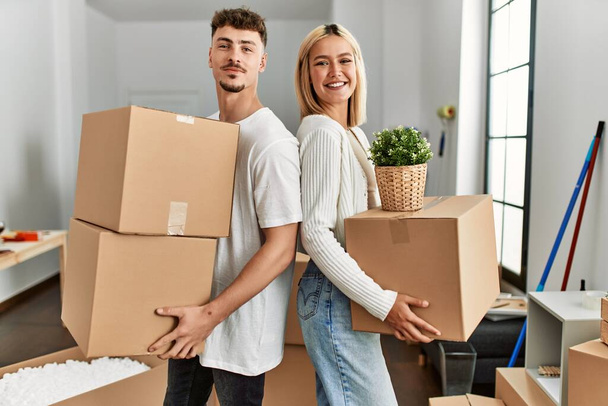 Νεαρό καυκάσιο ζευγάρι χαμογελά χαρούμενος κρατώντας κουτιά από χαρτόνι στέκεται στο νέο σπίτι. - Φωτογραφία, εικόνα