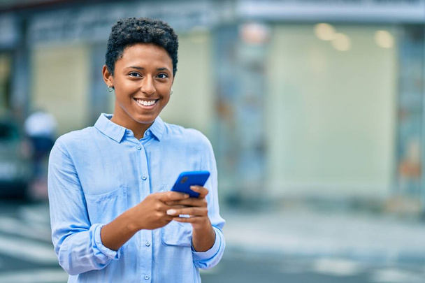 Genç Afrikalı Amerikalı kız şehirde akıllı telefon kullanarak mutlu bir şekilde gülümsüyor.. - Fotoğraf, Görsel