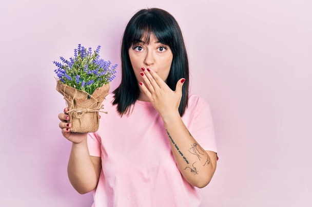 Jeune femme hispanique tenant une plante de lavande couvrant la bouche avec la main, choquée et effrayée par l'erreur. expression surprise  - Photo, image