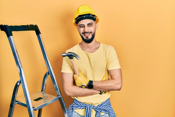 Handsome man with beard wearing hardhat holding hammer by stairs winking looking at the camera with sexy expression, cheerful and happy face.  - Photo, Image
